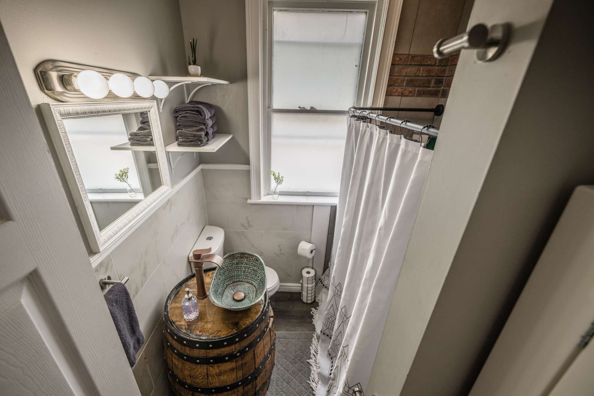 A compact bathroom with a toilet, sink, and a small window. There is a wooden barrel, a mirror, and a shelf with towels.