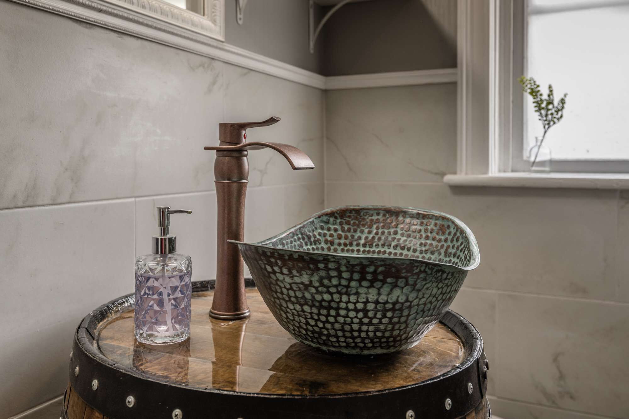A bathroom with a marble wall, a copper faucet, a glass soap dispenser, and a decorative bowl on a wooden barrel.