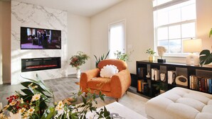 A living room with a marble fireplace, a television mounted on the wall, a comfortable orange armchair, a white sofa, a bookshelf, and a large window with natural light. - ~Weekly 15% OFF~ Modern Charm w/ Luxury Beds, Fireplace, Workspace (Luray)