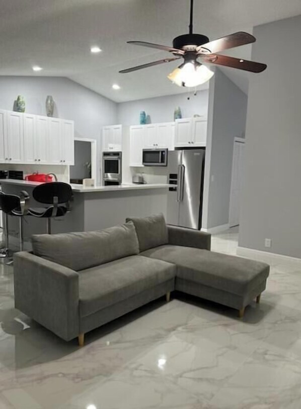 A modern living room with a grey sectional sofa, a ceiling fan, and a kitchen area in the background.
