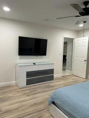 A bedroom with a bed, a TV mounted on the wall, a ceiling fan, and a door leading to another room.