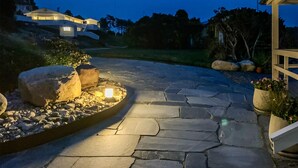 Een pad van stenen leidt naar een huis met een verlichte tuin, grote rotsen en potplanten.