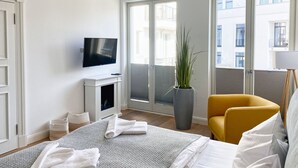 Ett hotellrum med en säng, en tv, en krukväxt, en gul fåtölj och en balkong med utsikt över byggnader.