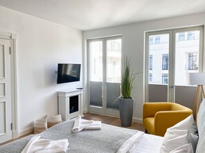 Camera d'albergo con un letto, una televisione, una pianta in vaso, una poltrona gialla e un balcone con vista sugli edifici.