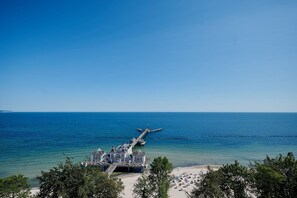 A coastal hotel with a pier extending into the sea, a beach area with sun loungers, and a clear view of the ocean. - Pool und Sauna mit Meerblick, Direkt an der Seebrücke - First Sellin Fewo 3.8 - First Sellin (Sellin)