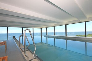 Una piscina interna moderna con pavimento in vetro, ampie finestre che offrono vista sul mare e una scala metallica per accedervi.