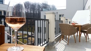 A balcony with a glass of wine, wicker chairs, and a view of a building and trees. - Pool und Sauna mit Meerblick, Direkt an der Seebrücke - First Sellin Fewo 3.8 - First Sellin (Sellin)