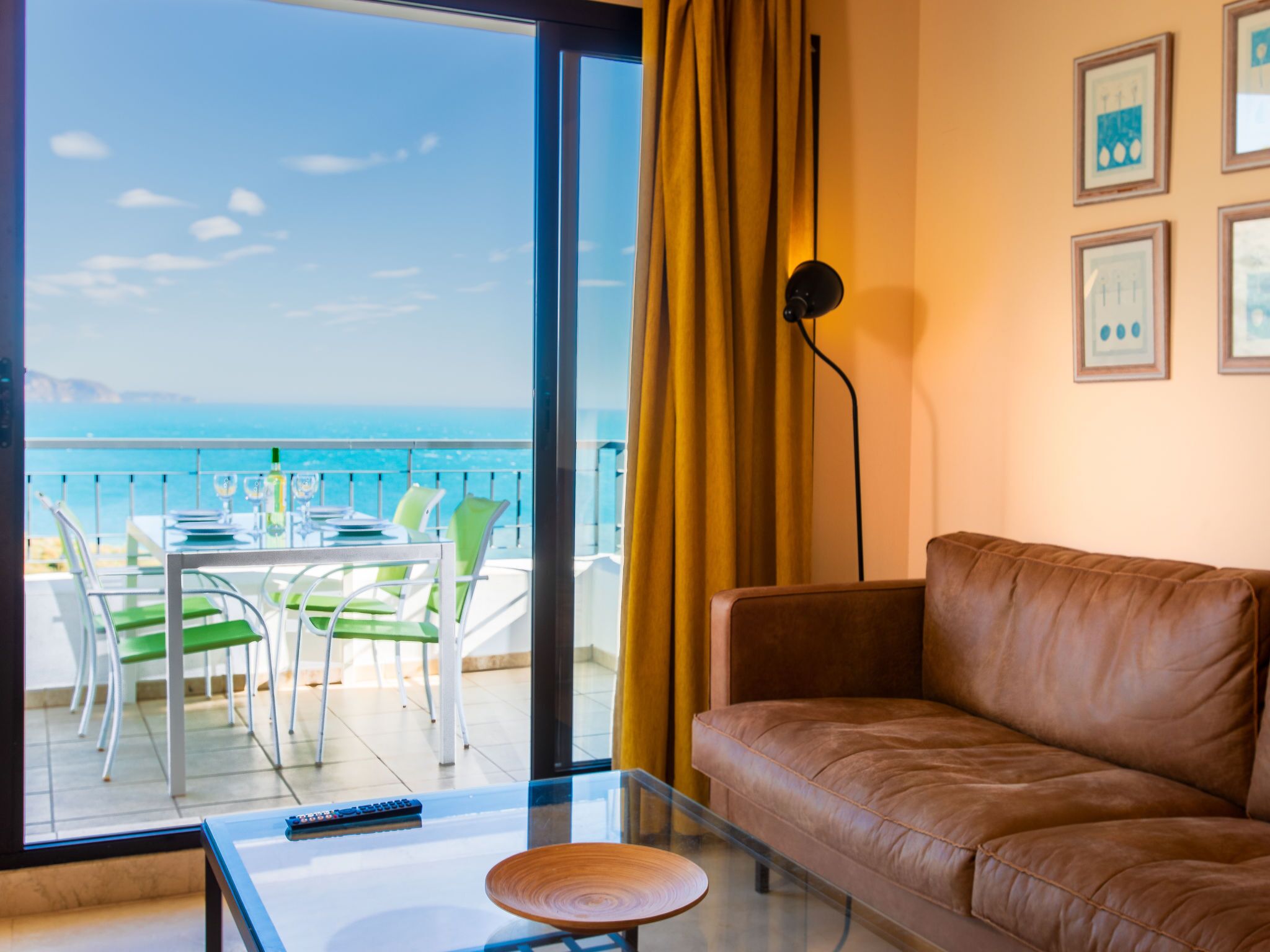 A room with a leather sofa, a glass coffee table, a remote control, and a view of the ocean through large windows.