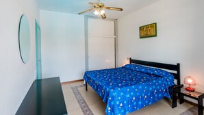 Quarto com cama, escrivaninha, ventoinha no teto, mesa de cabeceira e quadro na parede.