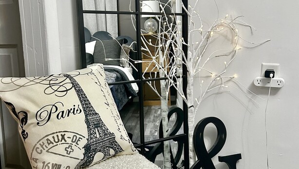 Ein Zimmer mit weißem, strukturiertem Hocker, einem Zierkissen mit dem Eiffelturm, einem schwarz-weißen Teppich und einer wandmontierten Leuchte.