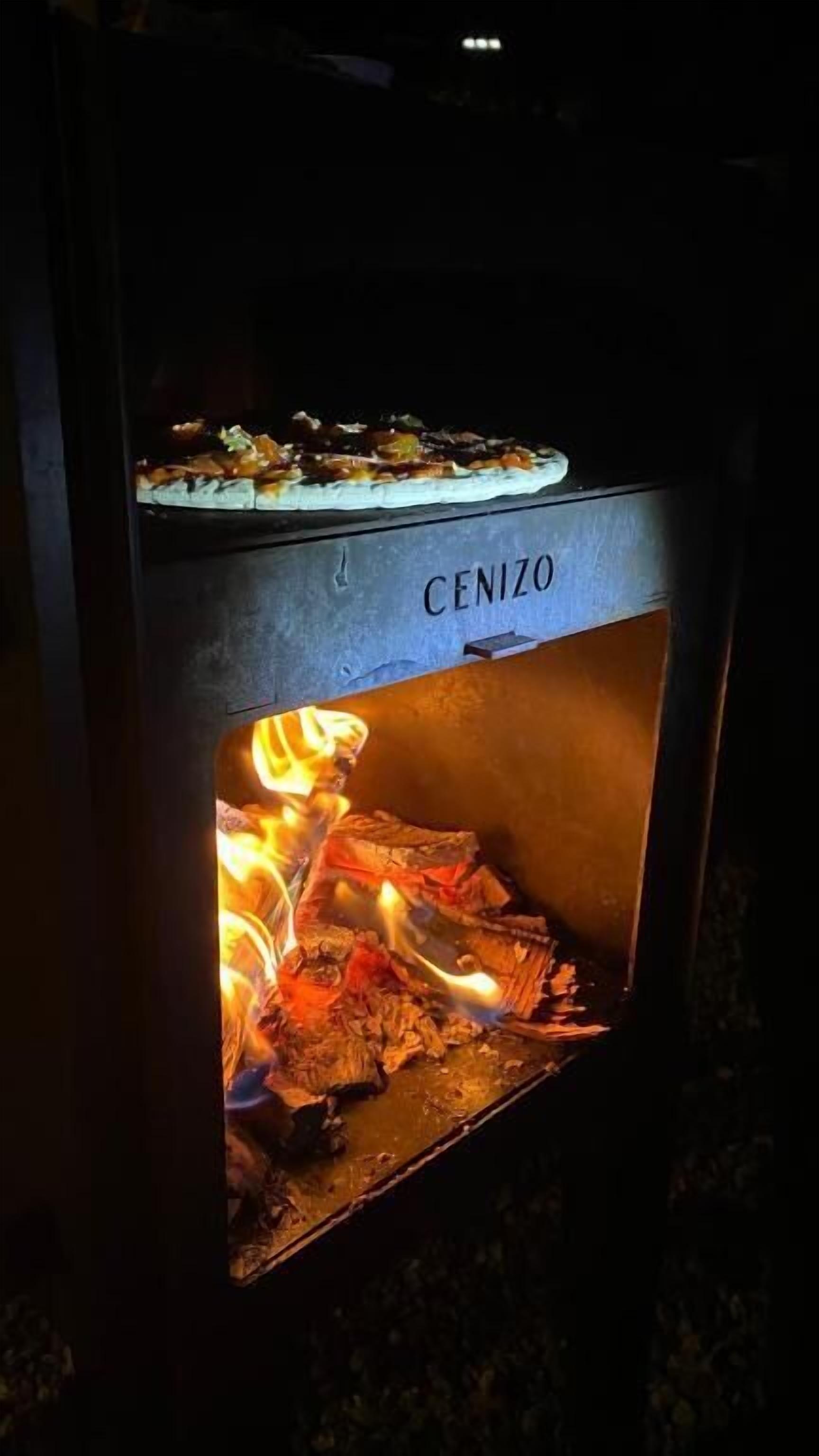 A wood-burning stove with a pizza on top and the word "CENIZO" on the front.
