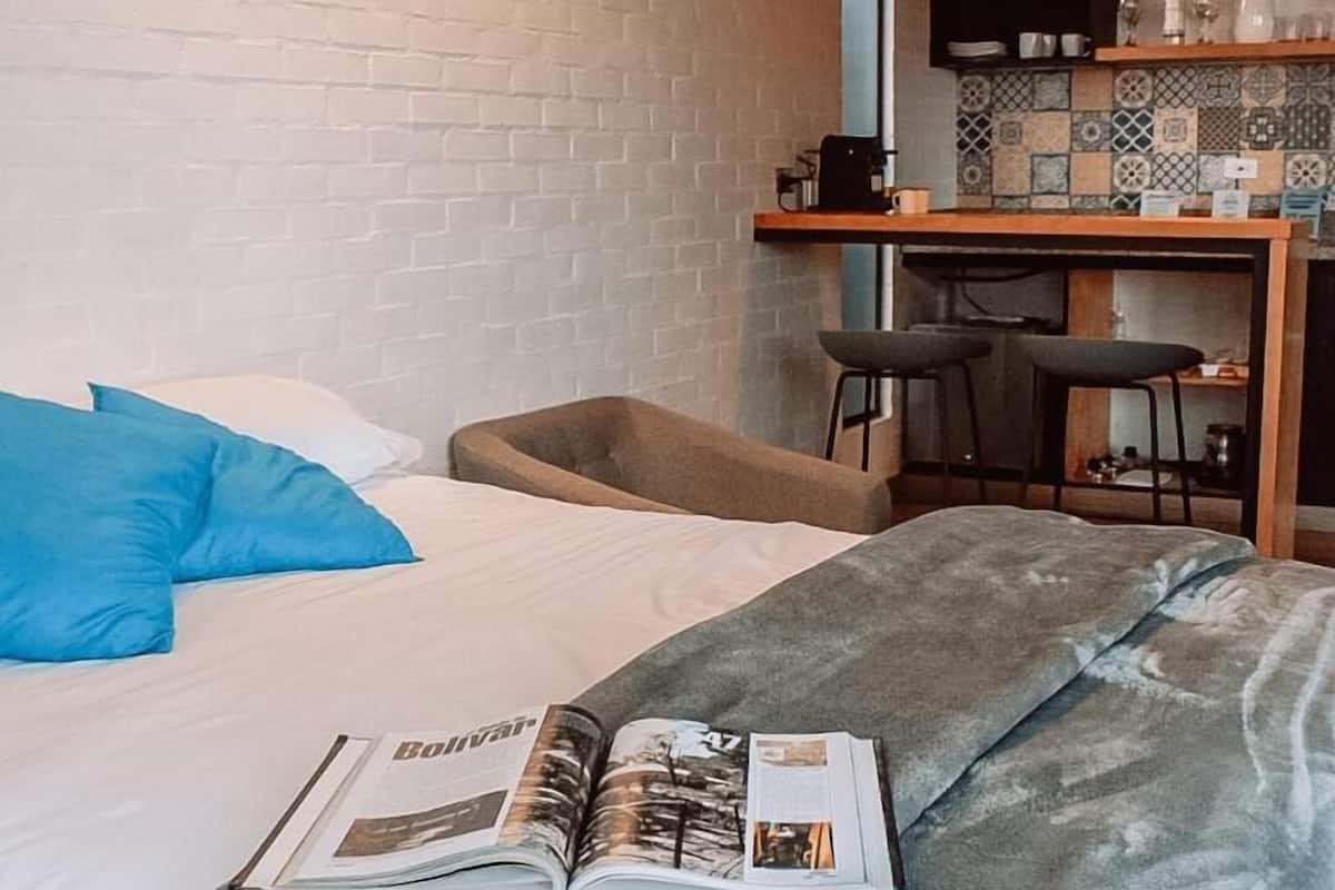 A hotel room with a bed, a reading area with a magazine, a kitchenette, and a TV.