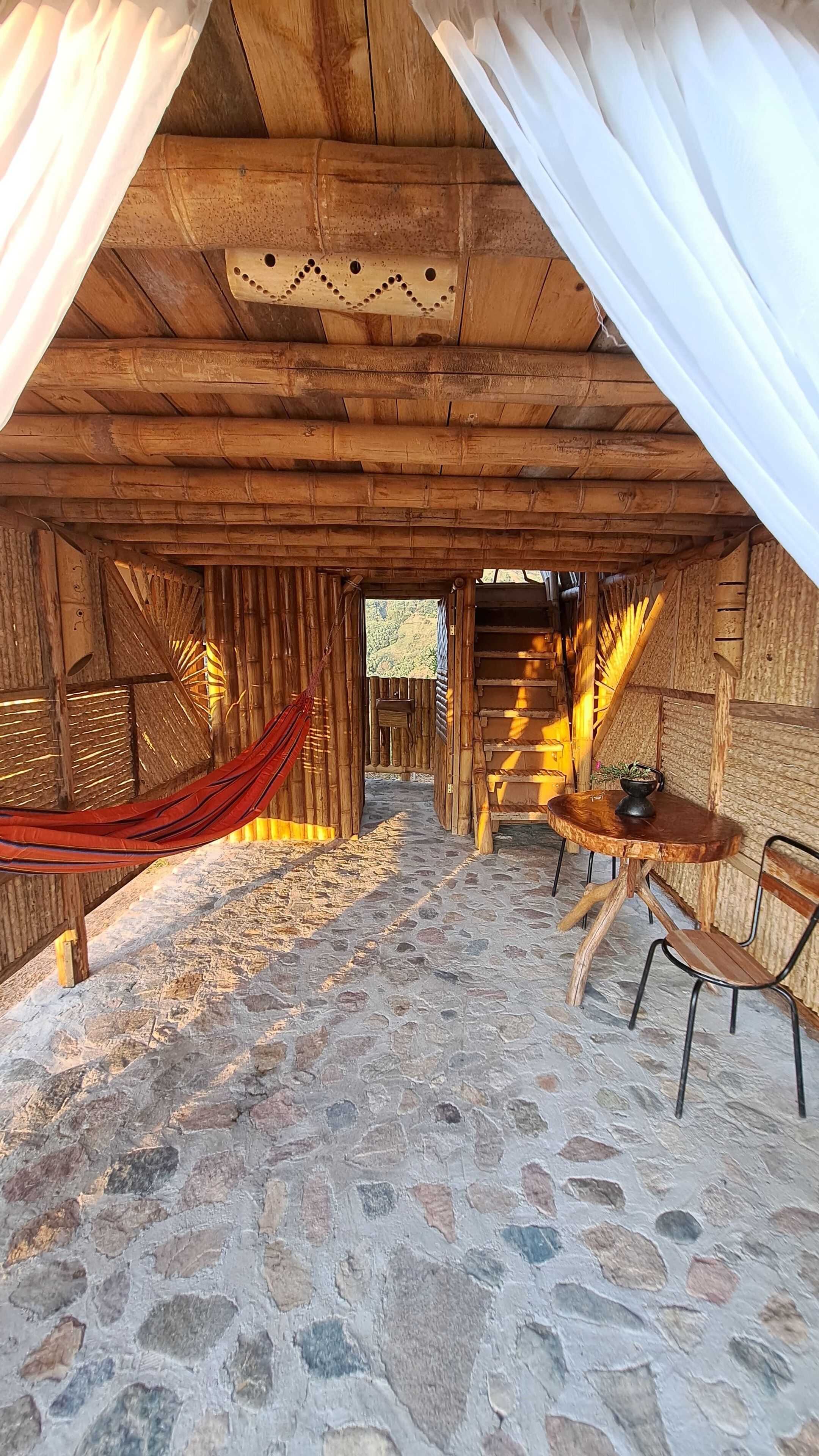 Um quarto rústico de madeira com soalho de pedra, uma rede, uma mesa de madeira com uma cadeira e uma escada que dá para uma área externa.