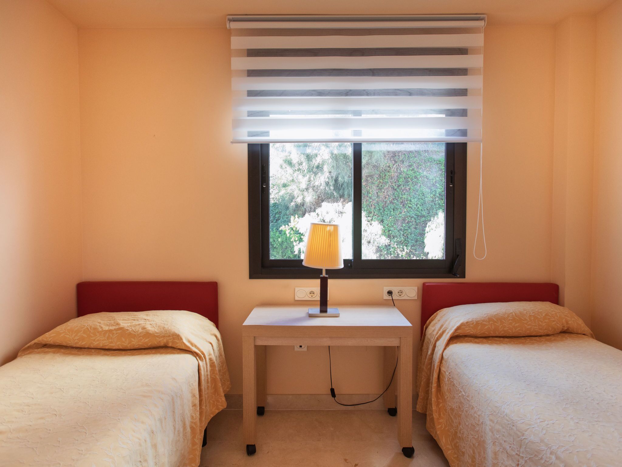 Een kamer met twee bedden, een bureau, een lamp en een raam met zonwering.
