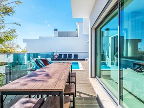 Ein moderner Poolbereich im Freien mit einem Holztisch und Liegestühlen, einer Glasreling und freiem Blick zum Himmel.