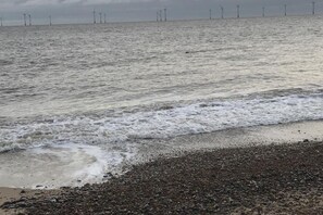 Une plage avec des vagues, des galets et des éoliennes visibles au loin.