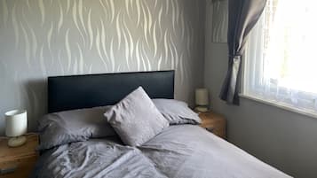 A bedroom with a patterned wall, a bed with grey bedding, a bedside lamp, and a window with curtains.