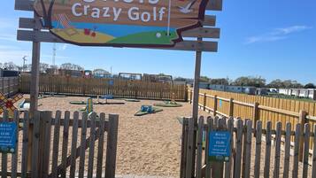 A wooden sign for ASIS Crazy Golf with a sandy play area in the background.