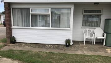 A single-story building with a white exterior, two windows with curtains, a small porch area with two white plastic chairs, and a green trash can.