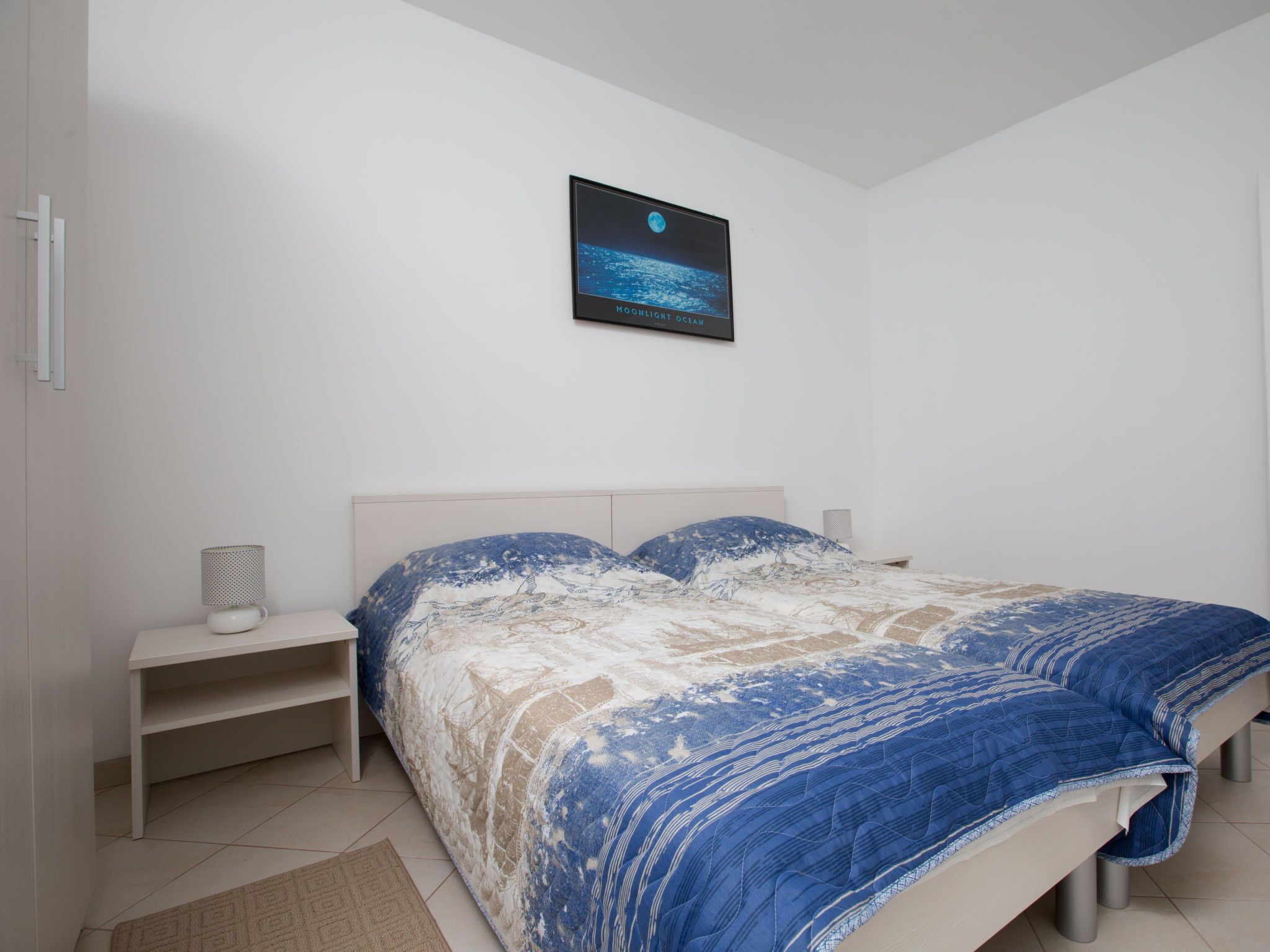 A bedroom with a bed, a nightstand, a wall-mounted TV, and a small shelf.