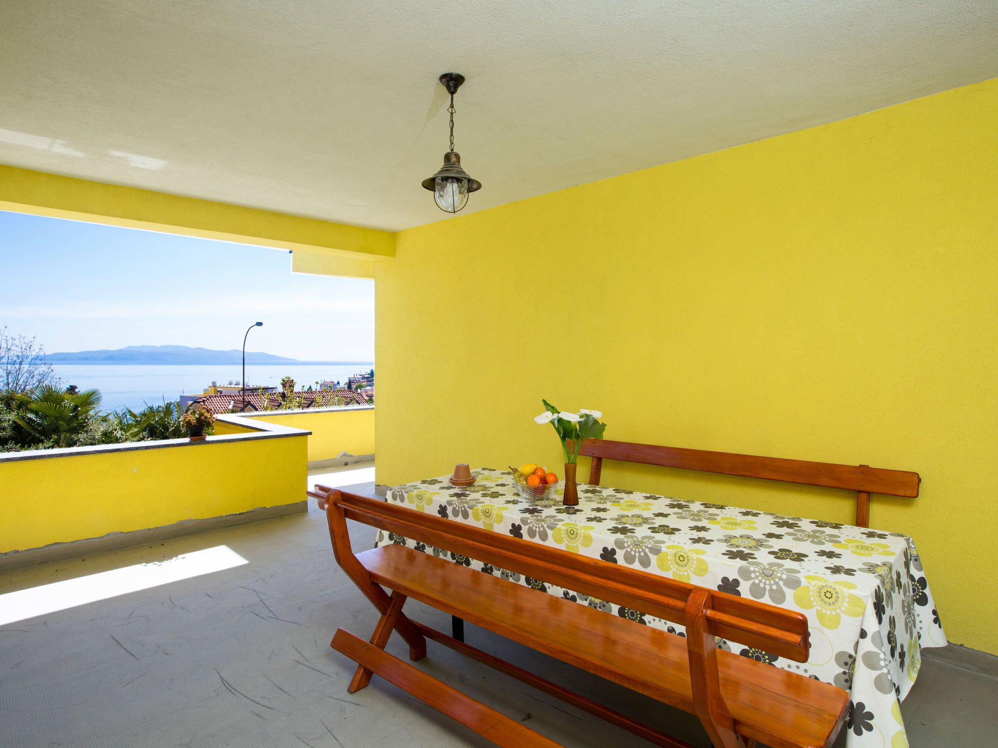 A room with a yellow wall, a wooden bench, a table with a floral tablecloth, and a view of the sea.