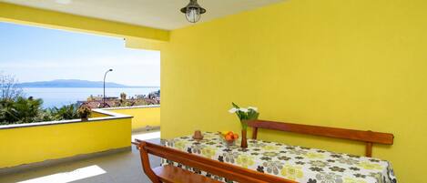 A room with a yellow wall, a wooden bench, a table with a floral tablecloth, and a view of the sea.