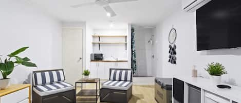 A modern, well-lit room with a bed, two striped armchairs, a small table, a ceiling fan, a TV, and a shower area.