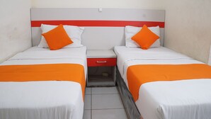 A hotel room with two beds, each with white bedding and orange accents, a red bedside table, and a small drawer.