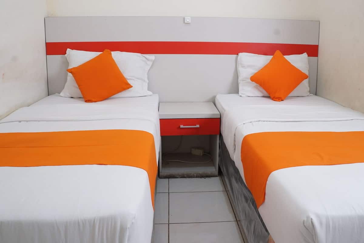 A hotel room with two beds, each with white bedding and orange accents, a red bedside table, and a small drawer.