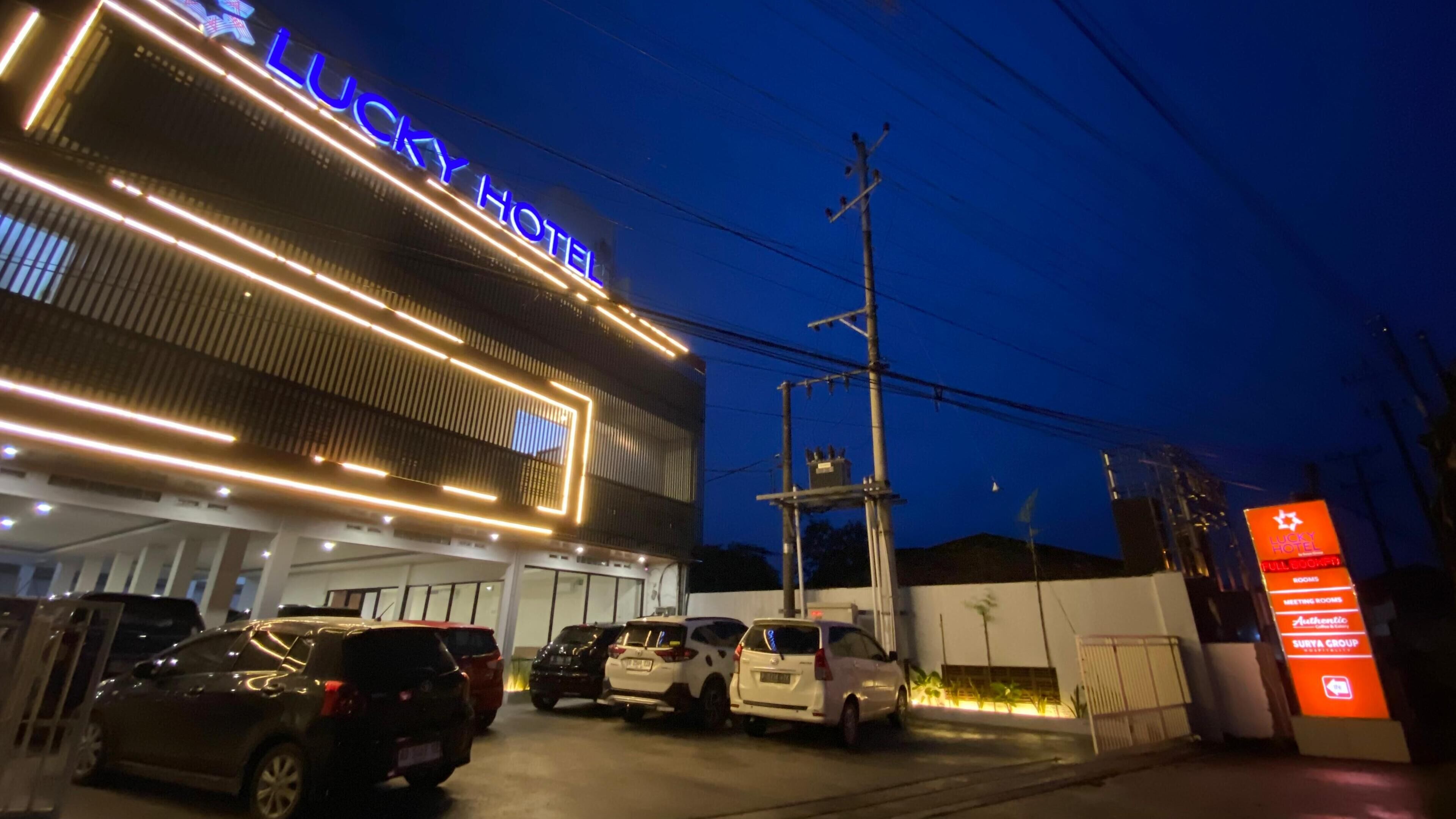 Un hôtel avec une enseigne bien visible, un parking où sont garées plusieurs voitures et une entrée fermée par un portail.
