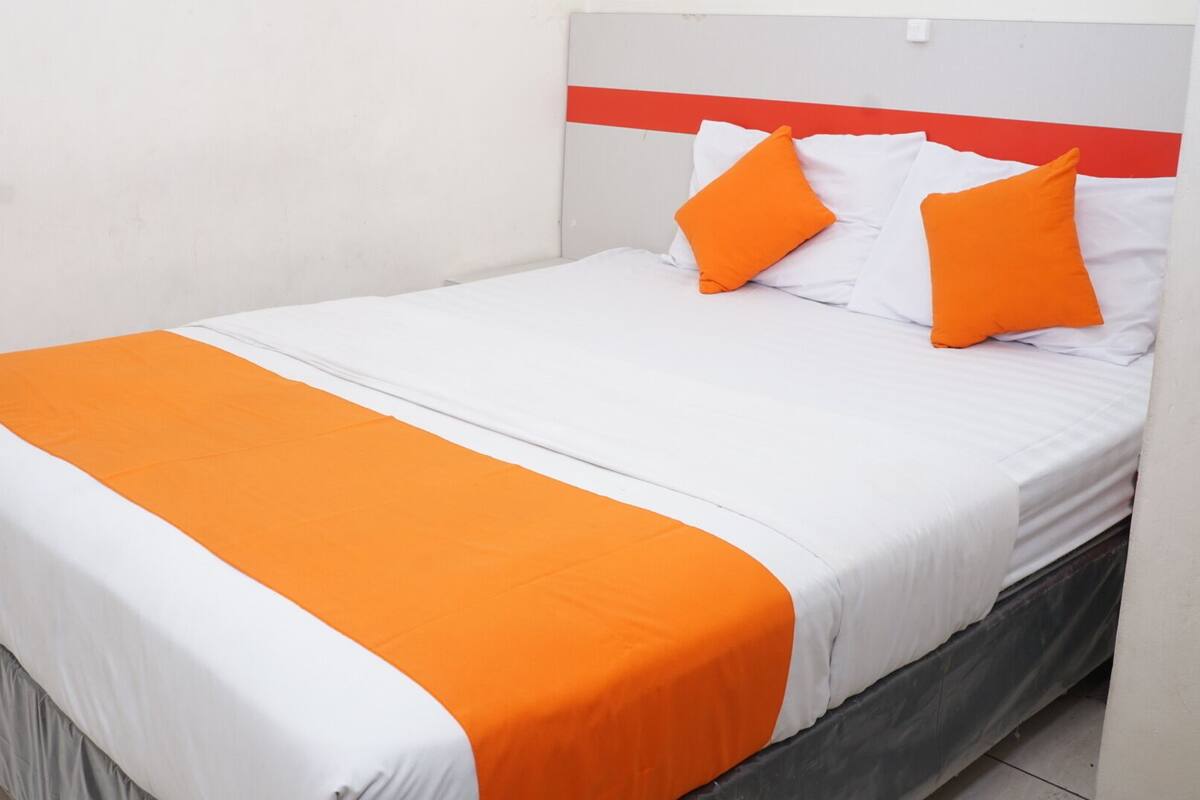 A single bed with a white and orange bedspread, two orange pillows, and a grey headboard with a red stripe.