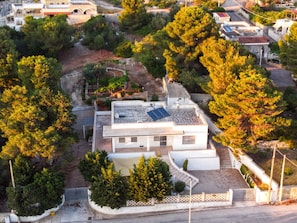 A modern two-story house with a flat roof, solar panels, and a fenced yard with trees.