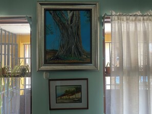 A framed painting of a tree on a wall, a smaller framed picture below it, and a window with a curtain.
