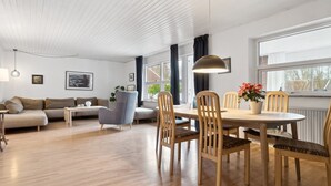 A modern living room with a wooden floor, a sofa, a dining table with chairs, and a large window with curtains.
