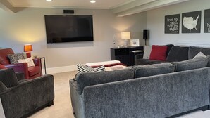 Ein modernes Wohnzimmer mit einem großen Flachbildfernseher, grauen Sofas, einem roten Sessel und einem kleinen Beistelltisch mit einer Lampe.