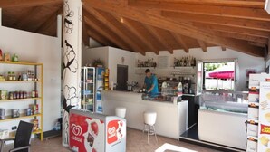 A café with a wooden ceiling, a counter with a person behind it, shelves with snacks, and a refrigerator.