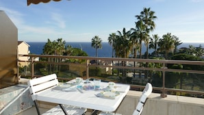 Un balcon avec une table dressée pour deux personnes, offrant une vue sur la côte, avec des palmiers et l’océan.