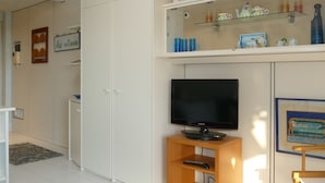 Un salon moderne avec un téléviseur à écran plat posé sur un meuble en bois, une armoire blanche et une étagère ornée d’objets décoratifs.