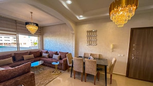 A modern living room with a sofa, a dining table with chairs, a chandelier, and a large window. - Luxury & Clean close to the Sea (Tangier)
