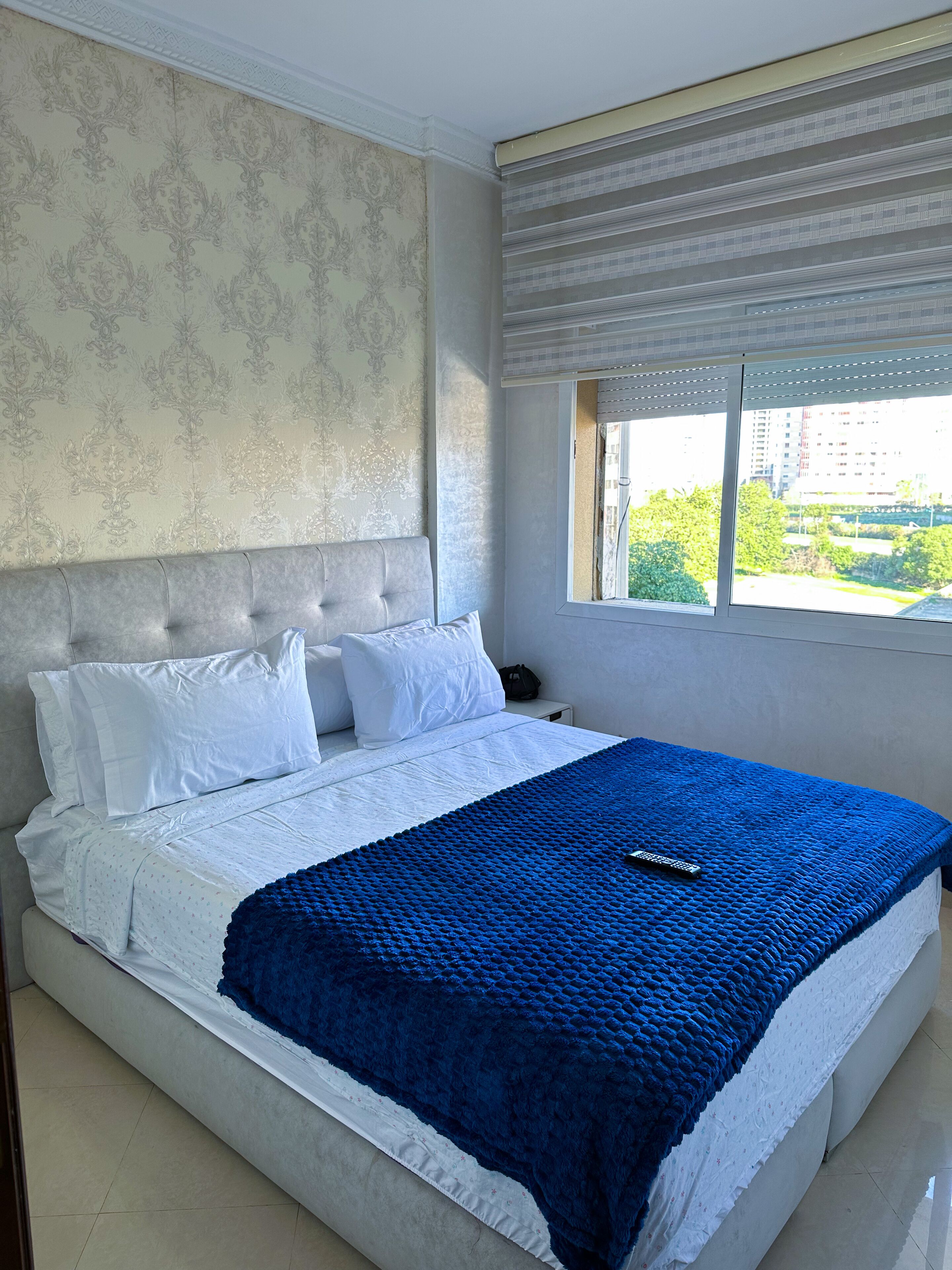 A neatly made bed with white and blue bedding, a headboard, and a window with a view of buildings and greenery.