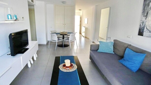 A modern living room with a flat-screen TV, a sofa with blue cushions, a dining area with a table and chairs, and a kitchen in the background. - El Grullo, with community pool. (Conil de la Fra.)