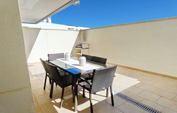 A rooftop patio with a table and chairs, a white wall, and a clear blue sky.