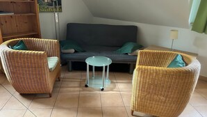 Um quarto com um sofá cinzento, duas cadeiras de vime, uma pequena mesa redonda e uma estante de livros.