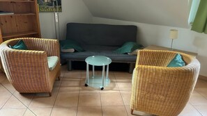 Una habitación con un sofá gris, dos sillas de mimbre, una mesita redonda y una estantería.