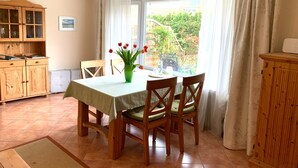 Een eetruimte met een tafel, stoelen, een groene vaas met bloemen, een houten kast en uitzicht door een raam.