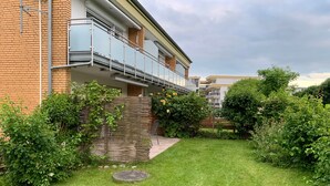 Een woongebouw met een bakstenen gevel, een balkon met glazen balustrade en een verzorgde tuin met diverse struiken en een gazon.