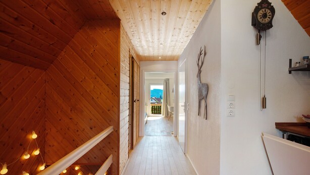 Un couloir intérieur en bois, avec un plafond, une horloge et une vue sur l’extérieur par une porte ouverte.