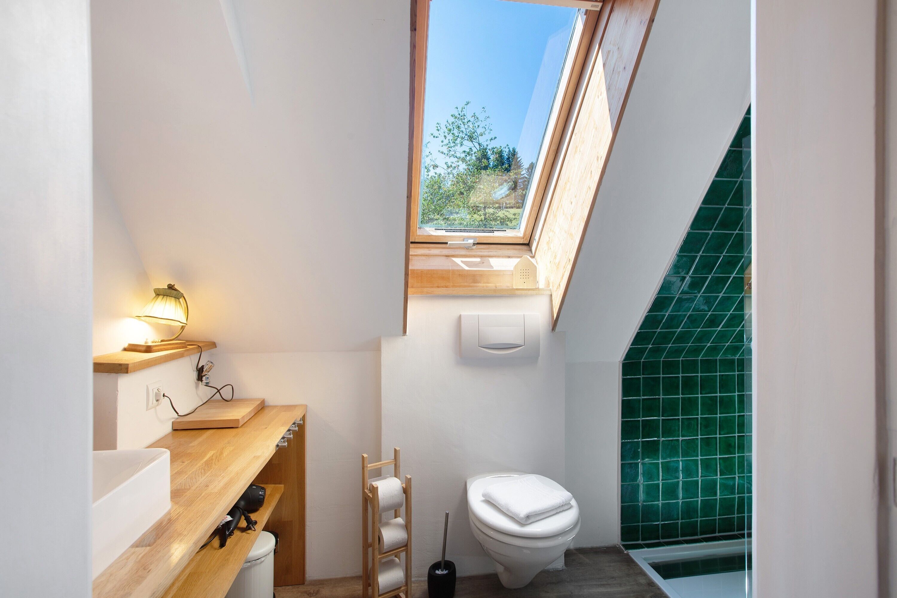 Ein kleines Badezimmer mit Oberlicht, einem Holzbadezimmerhocker, einer Toilette und einer grün gefliesten Duschkabine.