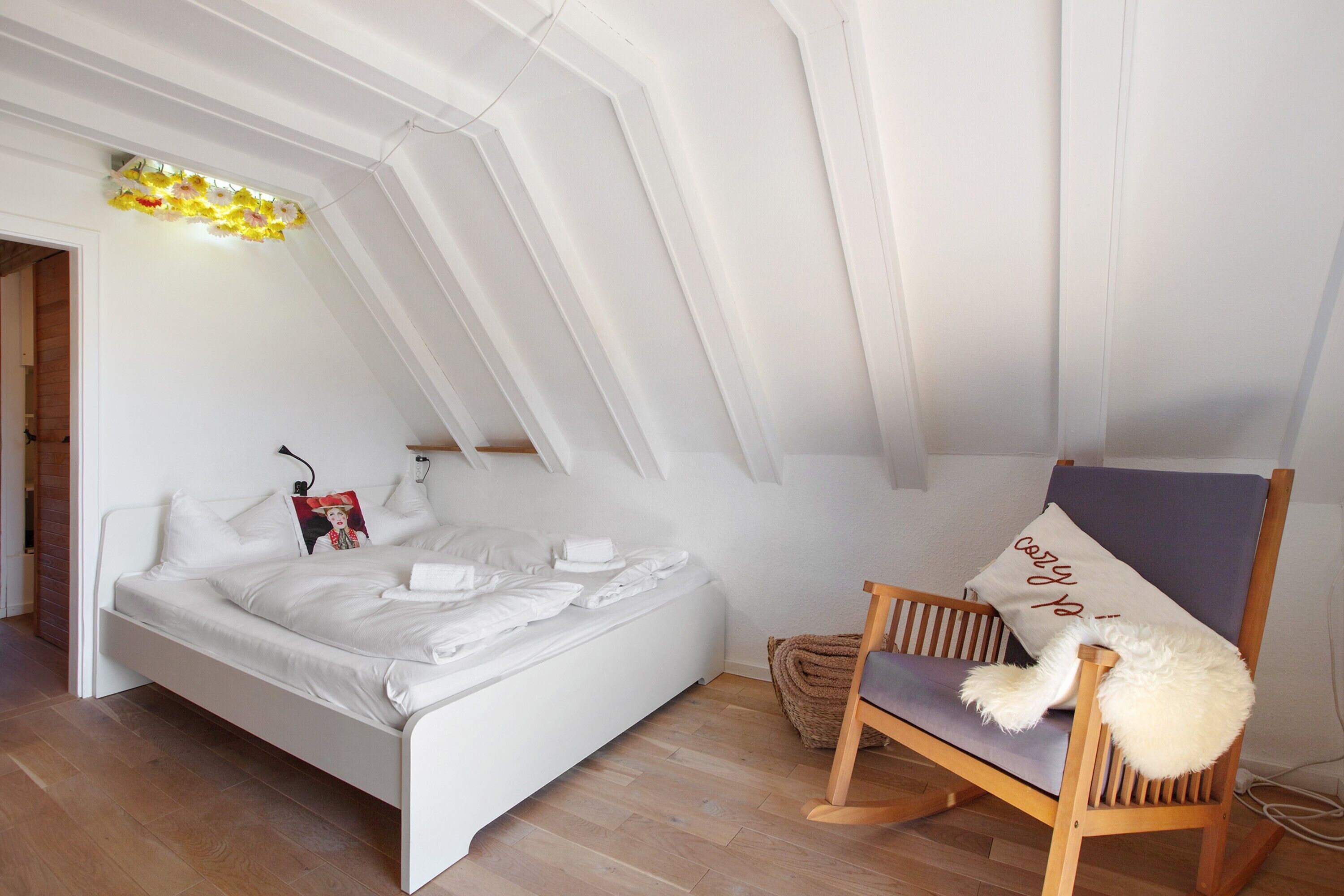 Une chambre avec un lit blanc, une chaise en bois, un coussin orné d’un motif, et un petit panier posé sur le sol.