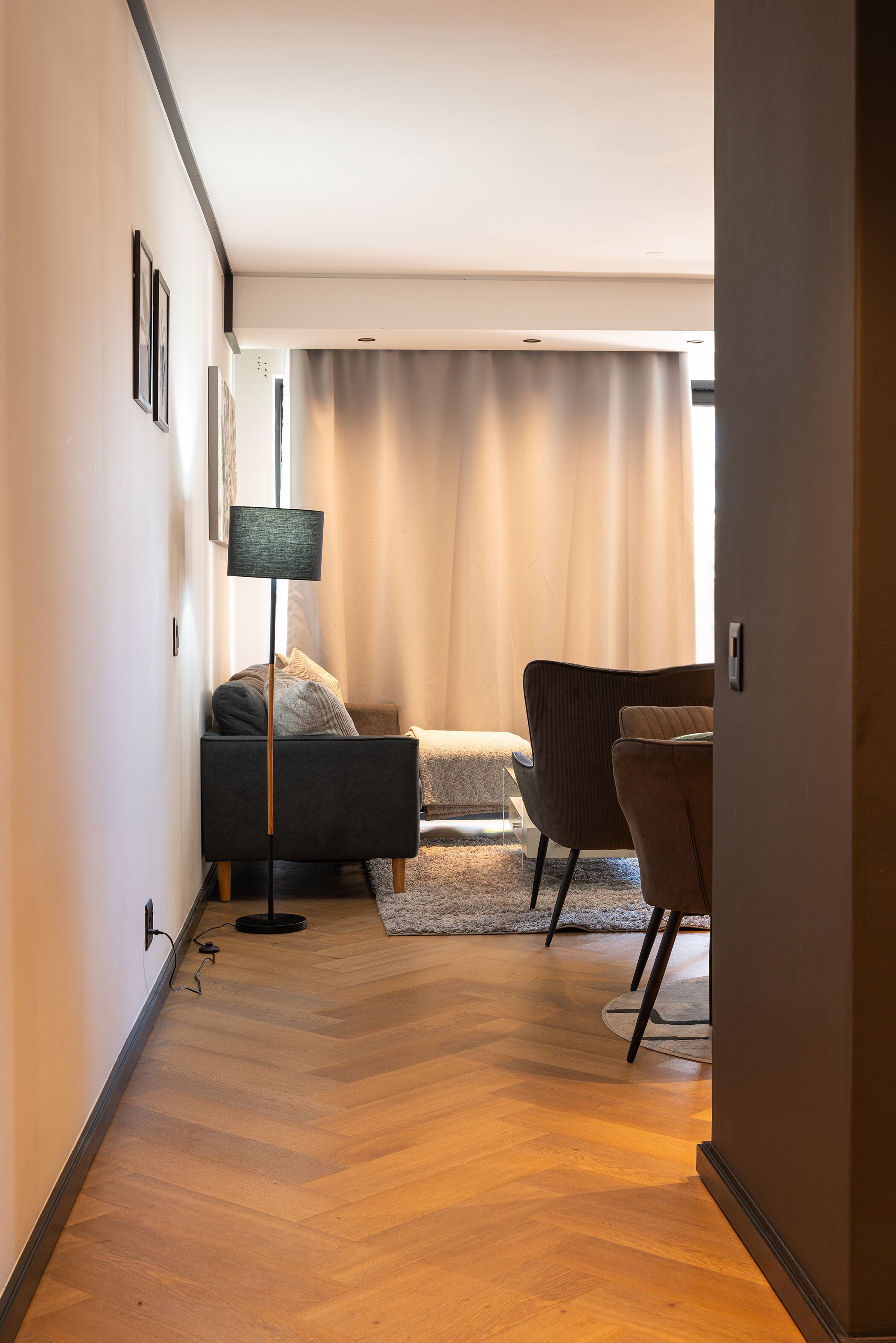 Et moderne hotelværelse med trægulv, en seng med mørkt sengegavl, en grå lænestol, en gulvlampe og et stort vindue med gardiner.